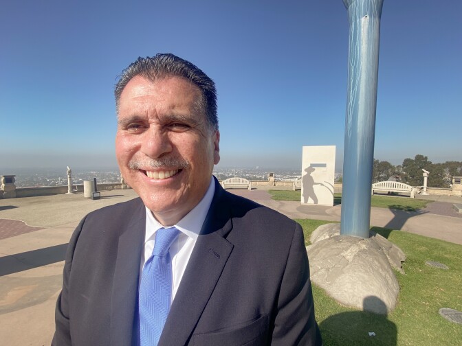 A man in a dark suit and blue tie smiles for the camera. Behind him we see a park and some sort of public monument.