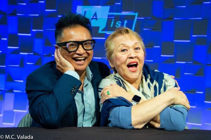 Alec Mapa, a dark-skinned man with black hair wearing a blazer and black glasses and Amy Hill, a white woman with short blond hair crossing her arms, smile at the camera