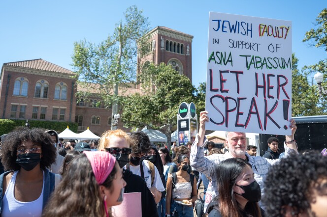 A group of people, mostly wearing masks, congregate around a man holding up a sign that reads, "Jewish Faculty In Support Of Asna Tabassum Let Her Speak!" A red brick building with beige windows is visible in the backdrop.