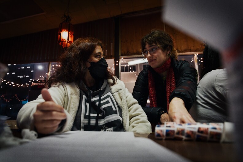 Two people sit at a table, while looking at each other. The person on the left is wearing a black face mask.