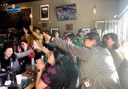 Smiling soccer fans clap and high-five each other while inside a restaurant. In the background, fans cheer at stadium on a television screen.