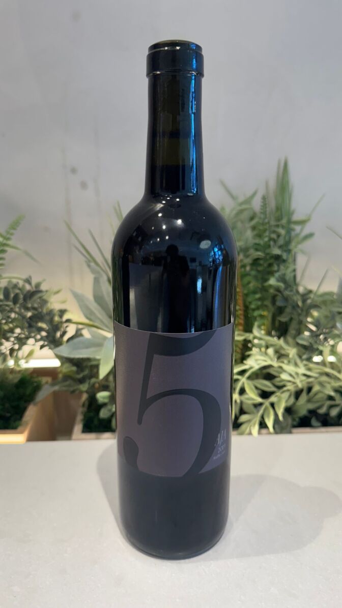 A bottle of red wine with the number 5 on the label on a white countertop with plants in the background. 