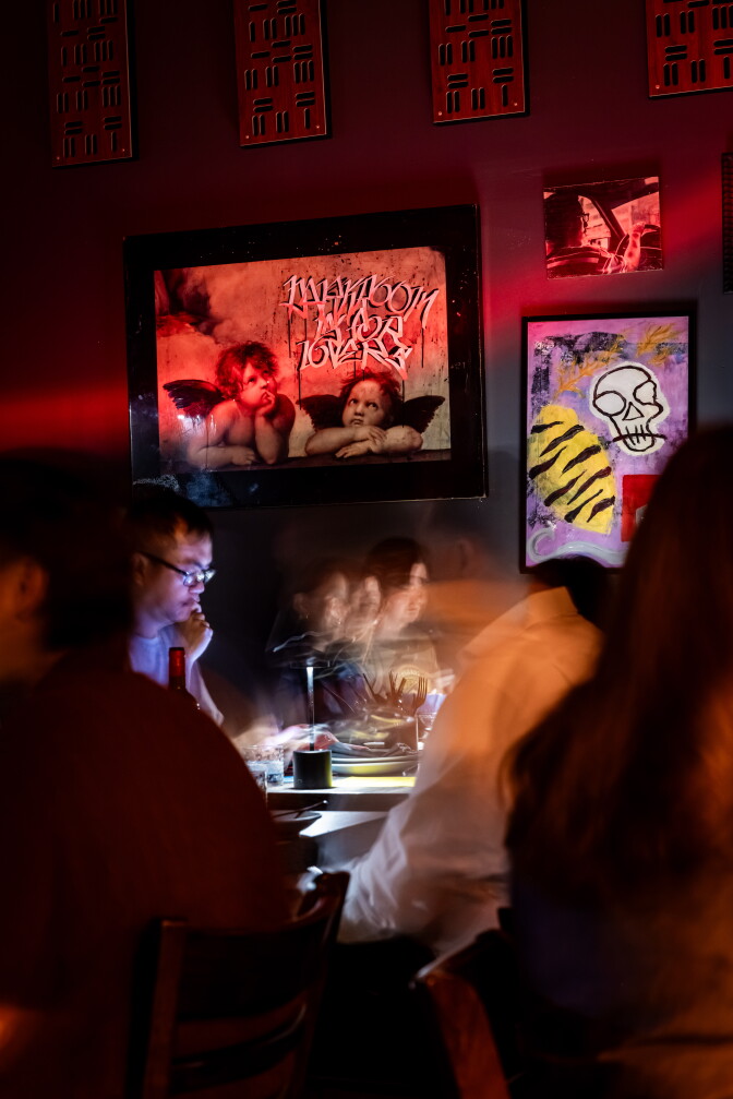 Dimly lit restaurant interior with red-toned lighting, framed artwork including cherubs with graffiti text and a skull painting, and blurred diners at a table in the foreground.