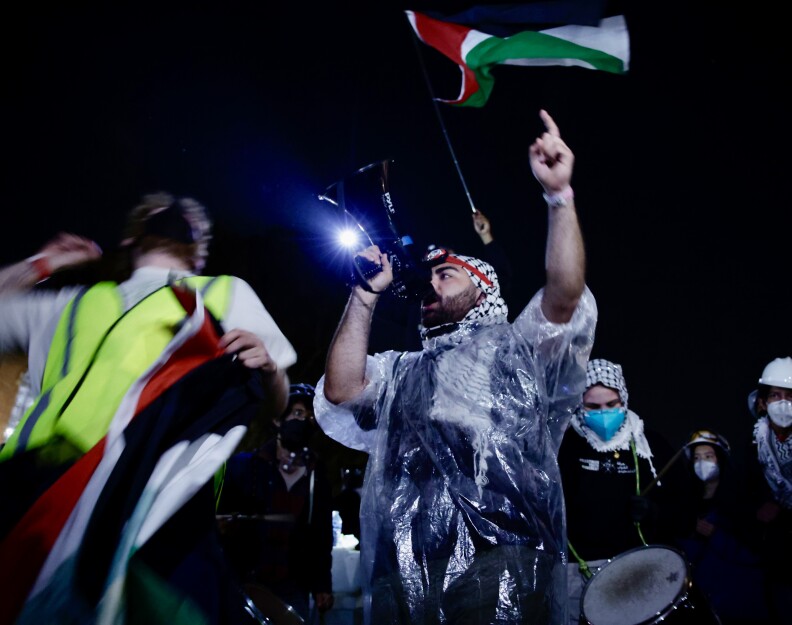 A pro-Palestinian protester wears a thin plastic rain coat and carries a megaphone. He wears a Keffiyeh and a Palestinian flag is being waved behind him by another person.