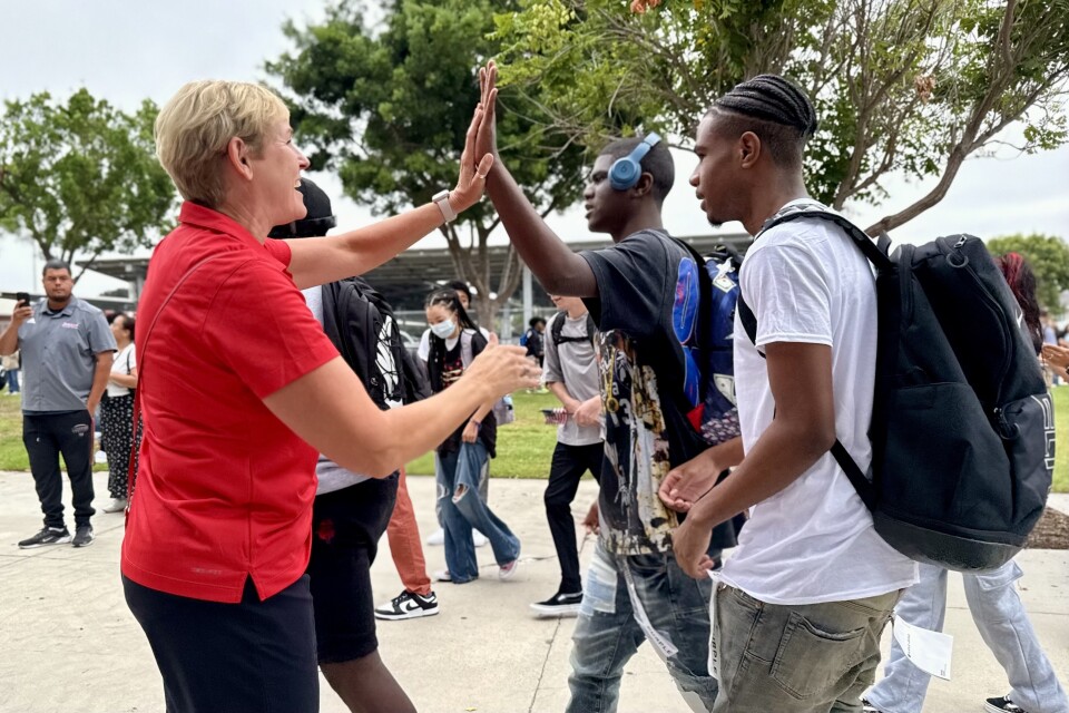 A woman with light skin tone wearing a red polo high fives a teen boy with dark skin tone and blue headphones, as other people walk by outside.