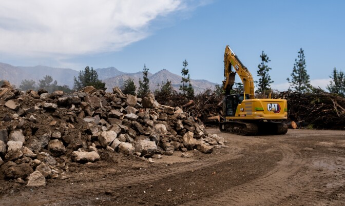 A large pile of concrete with trees in the background. A big yellow machine is seen on the far end of the pile.