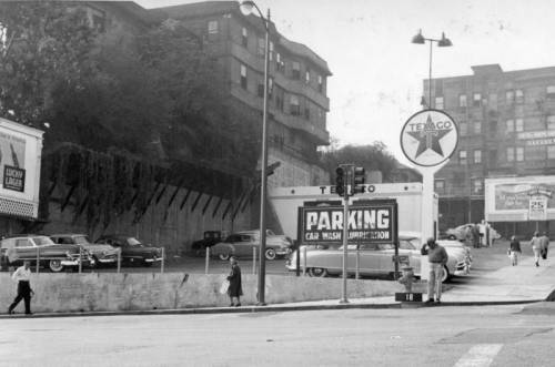 The photo is a black and white image of an urban street scene, likely from a past era based on the architecture and vehicles. A multi-story apartment building is on the left, and another large structure is on the right. There's a Texaco Sign prominently displayed in the center with the recognizable star logo. Below the Texaco sign, there's a lot with signage reading "PARKING CAR WASH LUBRICATION." Several cars are parked there. People are walking along the sidewalk in front of the lot. On the left side, there's a billboard advertising "LONDON GUARANTEE & ACCIDENT COMPANY LIMITED."