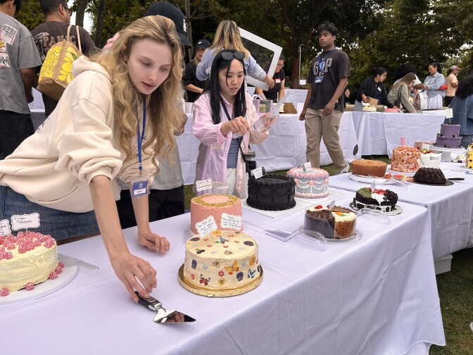 Two women are standing in front of a table with cakes. One is leaning in.
