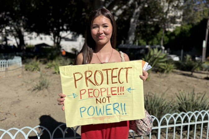 An adolescent girl with medium-light skin tone holds up a sign that says "Protect people!!! Not power!!!