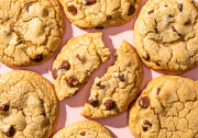 Several chocolate chip cookies on a light pink background. The cookie in the center is broken in half.
