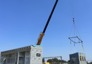 Students stand below a long crane machine, cradling the small portion of a house in pieces. The main portion of the house is at the front and tho the left of them. They are all wearing hard hats and safety vests.