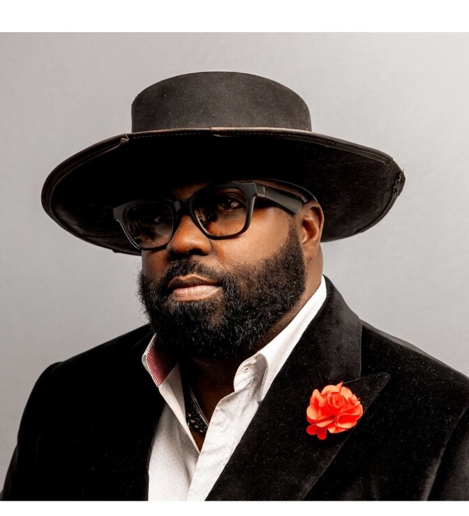 A black man with a short beard wearing a wide-brimmed black hat, black-rimmed a glasses and a velvet black suit with a red flower pinned to the left lapel in a portrait. 
