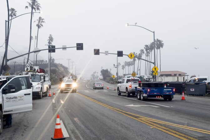 A highway with few cars on it. A truck approaches the camera with lens flares from the headlights. Another truck drives away from the camera. To the left of frame more work trucks line the shoulder. It's foggy and not yet bright.