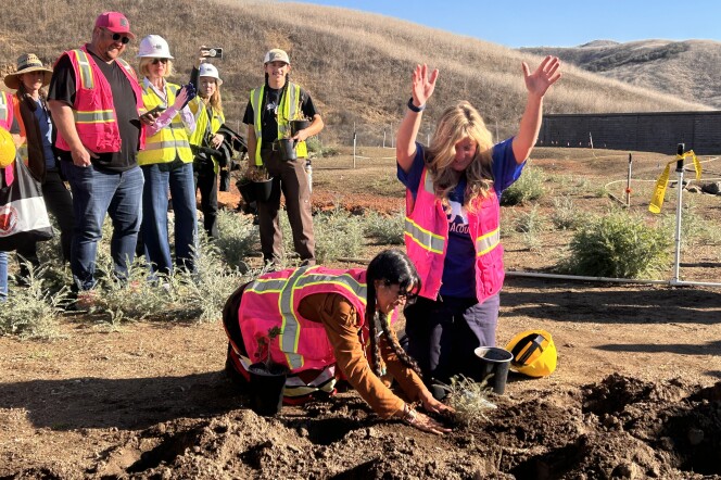 Two women are kneeling over a hole buried in the dirt ground around a small green plant. The woman on the left has long black hair tied in two braids and a bright pink high-visibility vest with her hands around the plant. The woman on the right has long blonde hair past her shoulders is wearing the same pink vest over a navy blue t-shirt, with both her arms raised above her head.
