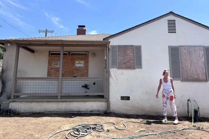 A person stands outside a house with boarded-up windows and a porch marked with 'No Trespassing' signs.
