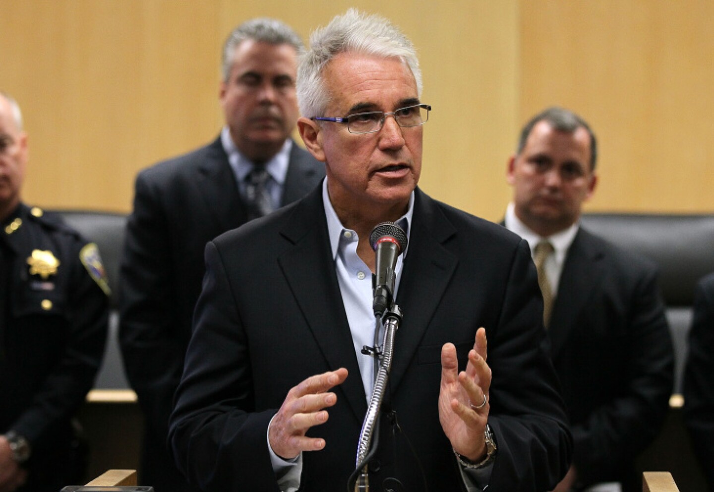 George Gascón, wearing a collared shirt and jacket but no tie, speaks in front of a microphone in what appears to be a courtroom.