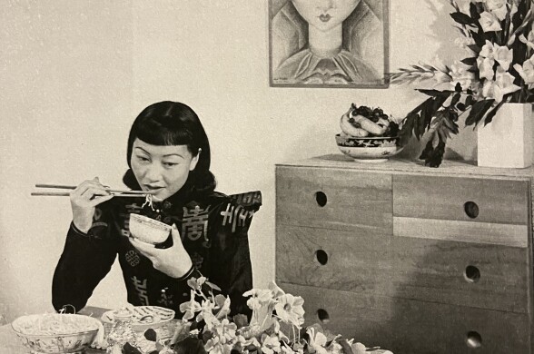 A sepia-toned photo of a Chinese American woman sitting in a mid-century modern dining room eating rice. 