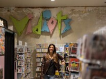 A feminine presenting person with light skin tone and light brown hair wearing a brown leather jacket and jeans stands leaning on a stand full of magazines. Behind them on the wall is a colorful sign that reads "Vidiots."