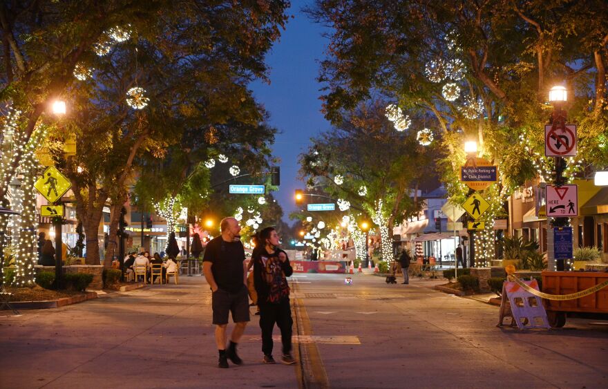 Two people walk in the middle of a closed-off street in the evening with trees and lights in the background.