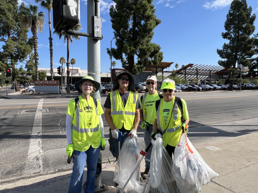 Four Volunteers Cleaning Communities members stand together for a picture. They all wear hats and neon yellow clothing. Three of them are holding trash bags.