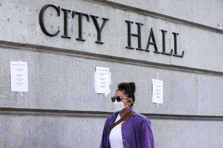 A woman wearing a purple jacket and a white face mask and sunglasses, with light brown skin, walks in front of a cement building with a sign reading CITY HALL. There are several pieces of paper posted to the wall.