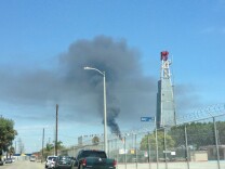 A oil drilling rig looms as smoke from a flare burns in the background of a residential street.