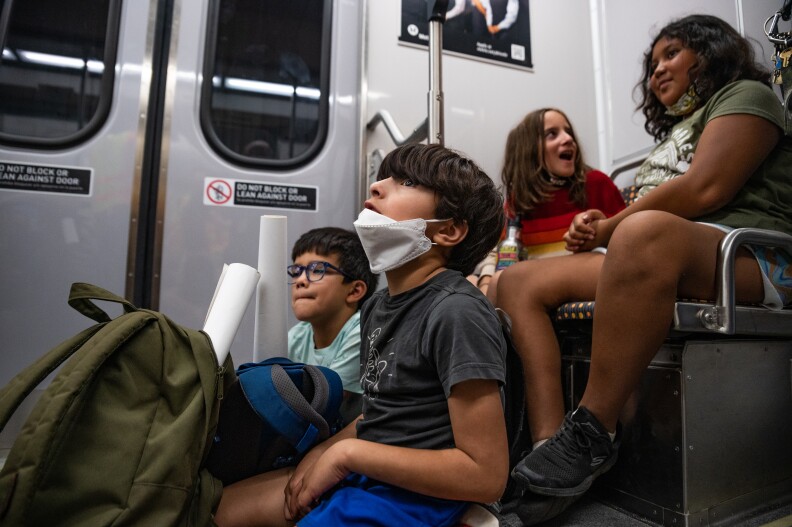 A group of children sit on the train. Two boys have backpacks on their laps, one with a mask hanging off his lower lip. Two girls sit behind them on a seat higher up, both smiling and one of them talking to the other.