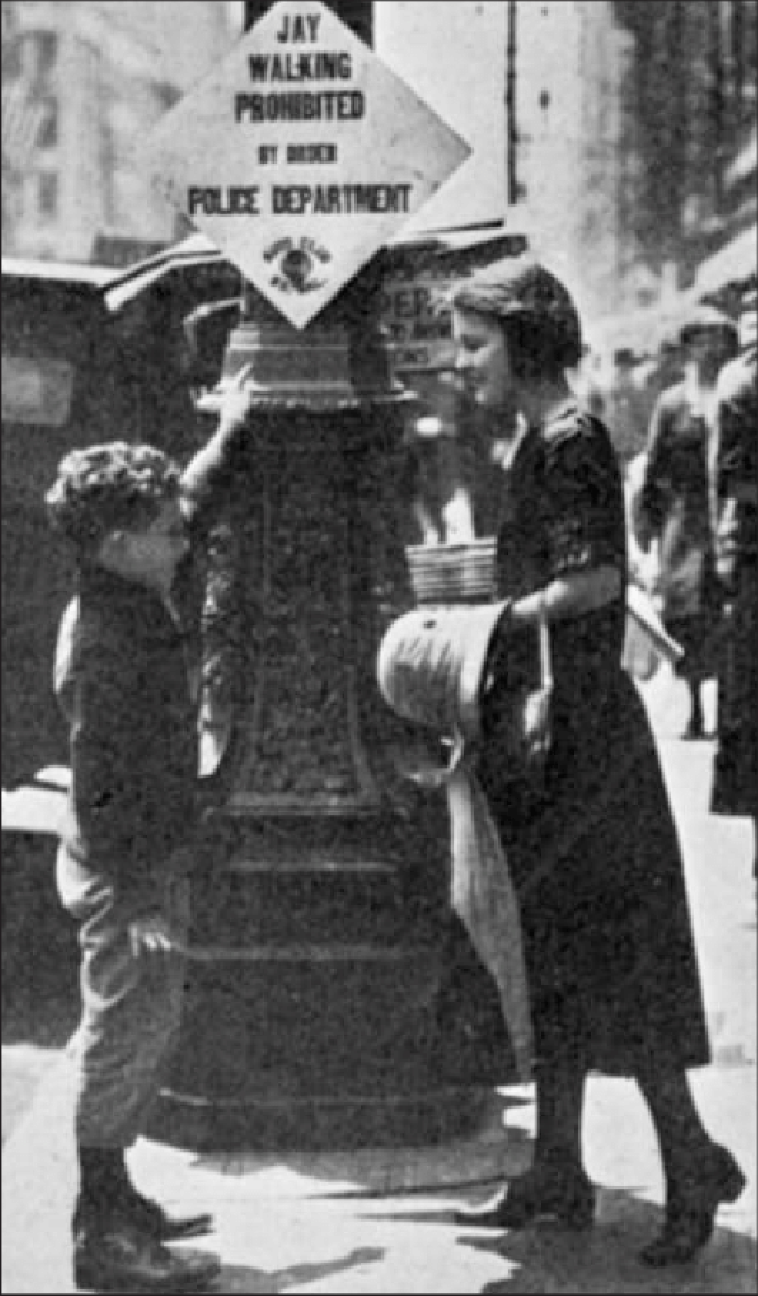 czarno-białe zdjęcie z 1923 roku przedstawia chłopca i młodą kobietę stojących przed znakiem drogowym z napisem "niedozwolone przechodzenie przez jezdnię" i " Wydział Policji."