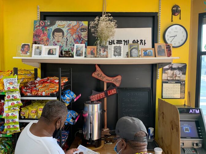 A scene from the inside of a vibrant convenience store, featuring two people getting coffee, as well as snacks, a clock, a sign in Korean, and about 10 small picture frames with portraits.