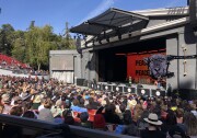 A large crowd fills the seats in front of a stage at an outdoor theater. The stage is decorated with a large illustration of a mountain lion with the words Peace Love beside it