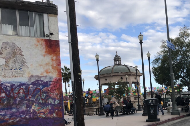 To the left a wall has a mural of a woman with graffiti over some landscape. To the right a park square features an elaborate  gazebo. People are walking through the park.