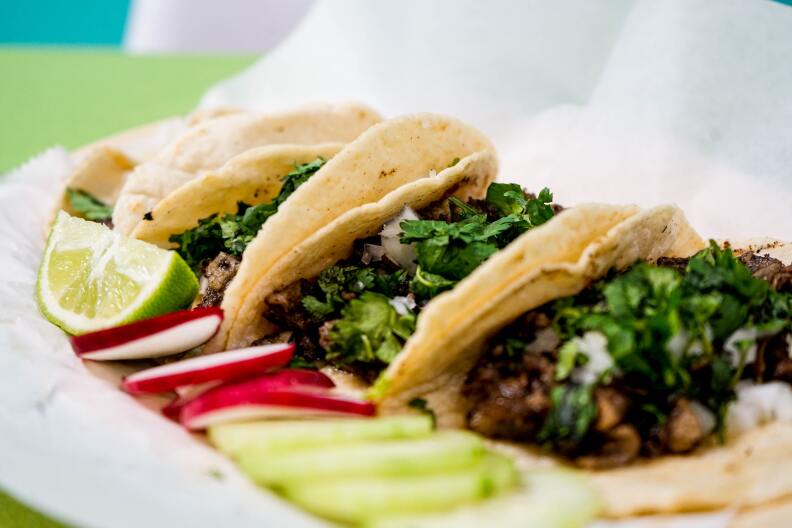 Three tacos in soft shells are lined up on a plate, leaning on one another. The meat in the tacos is browned, and all three have onions and cilantro on top of the meat. In front of the tacos on the plate is a lime, several slices of radishes and several slices of cucumbers.