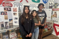 Three girls with varying skin tones stand in front of a series of tri-fold posters that are covered in photos and old news clippings.