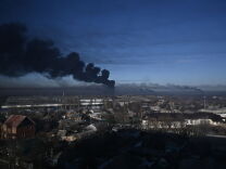 Black smoke rises above a military airport. Low buildings are seen in the lower half of the photo, a large shadow over the lower right portion of the photo. There is smoke rising from other areas further away. The sky remains largely blue with some light clouds, other than the black smoke.
