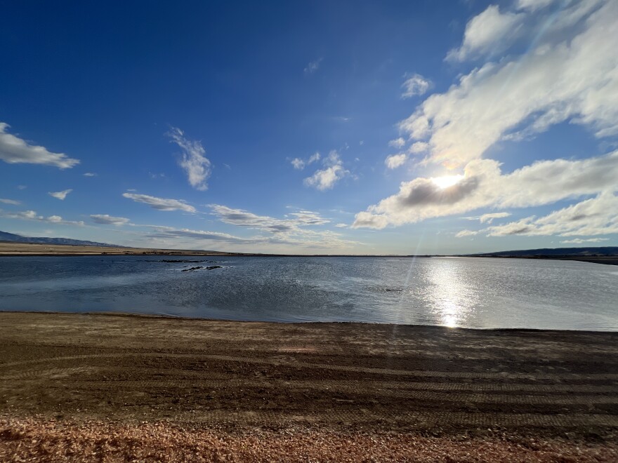 A large body of water separates a pale-blue sky with scattered clouds from dark muddy soil.