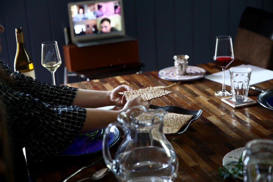 A woman's hands are seen breaking matzo bread at a table, which also contains wine, water, and other seder dinner elements in glasses and on plates. At the end of the table is a video screen with several squares showing others.