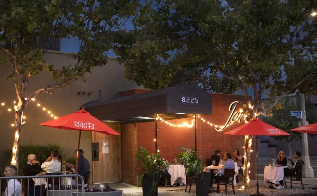 An image of the exterior of Jar, a restaurant located in the Beverly Grove neighborhood of Los Angeles.  The outdoor dining area consists of various tables and chairs underneath red umbrellas, all positioned around the restaurant's front entrance, which is covered by a long black awing next to a red brick wall.