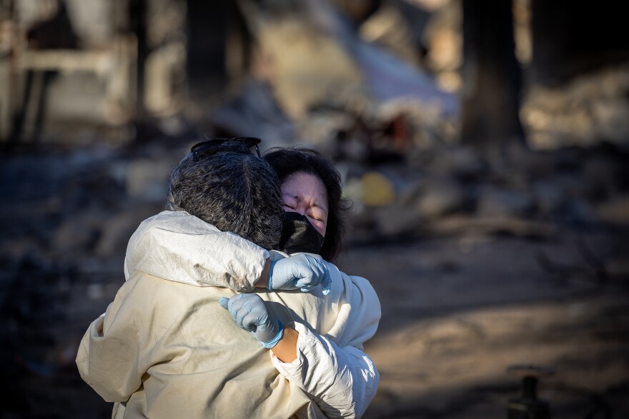A woman is wrapping her arms around another woman in a tight hug, her hands covered in blue latex gloves behind the other woman's back. The remnants of a burned home is out of focus in the background.