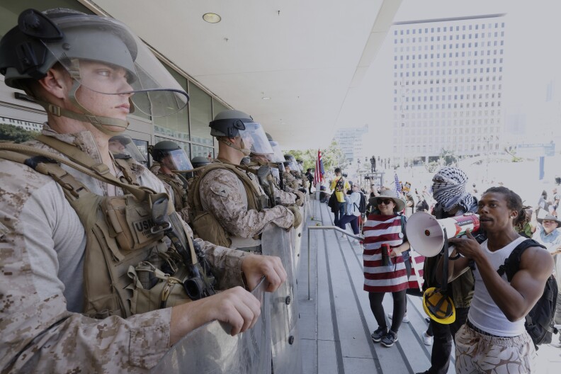 A row of troops hold shields outside a building as people gather on the steps. 