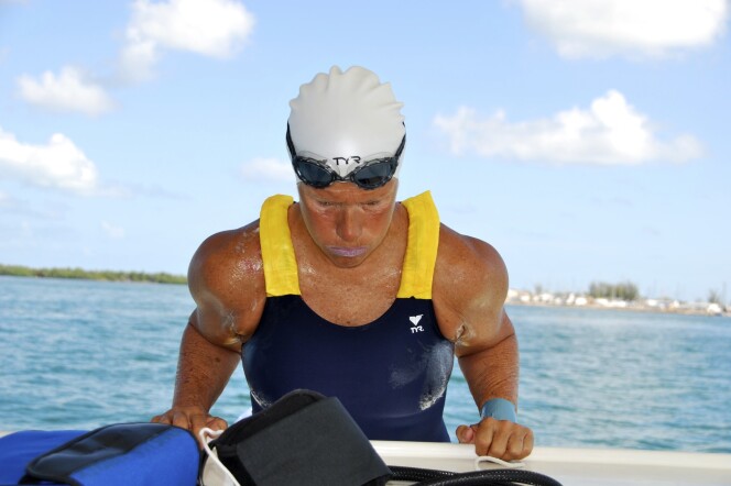Nyad is photographed while pulling herself out of the water into the side of a boat, elbows bent at her side. She wears a blue and yellow swim suit with a white swim cap and dark goggles above her brow. Her sunburned face looks downward, stoic with determination.