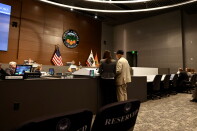 An older man wearing a beige vest and black hat stands next to a woman in a gray blazer as they stand behind a microphone and speak towards people sitting behind a gray dais. 