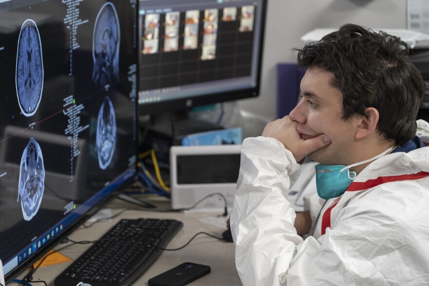A young white man, who appears to be a health care worker, is dressed in medical PPE and sits down examining brain scans on a large computer screen.