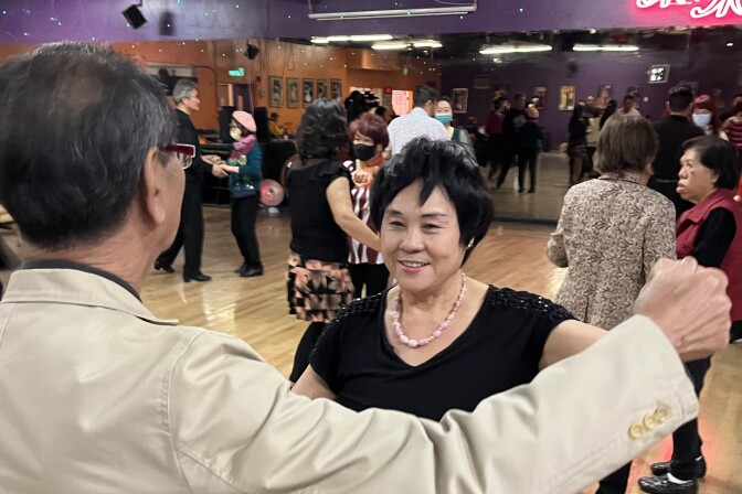 An Asian woman with short black hair dances on a busy dancefloor with an Asian man in a cream blazer with his back to the camera. 