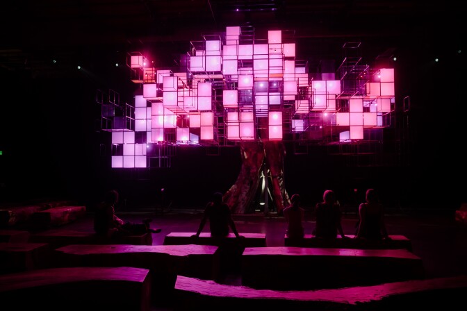 People sit in a dimly illuminated space, on wood benches, looking up at a faux tree. It's made from a real tree's trunk, with digital colored screens in square pixelated shapes creating a faux canopy. The panels at the time of this image are illuminated in a pink/purple color.