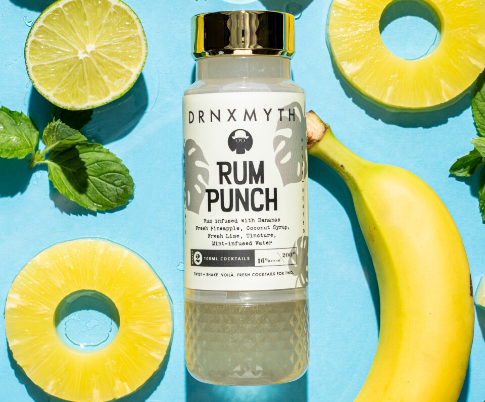 a bottle filled with pale liquid against a blue background surrounded by bananas and slices of pineapple and lime