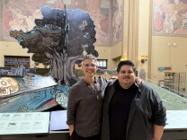 Two men pose in front of a giant pop-up-book art installation featuring a tree, a feathered serpent and a sea turtle inside the LA Central Library rotunda.
