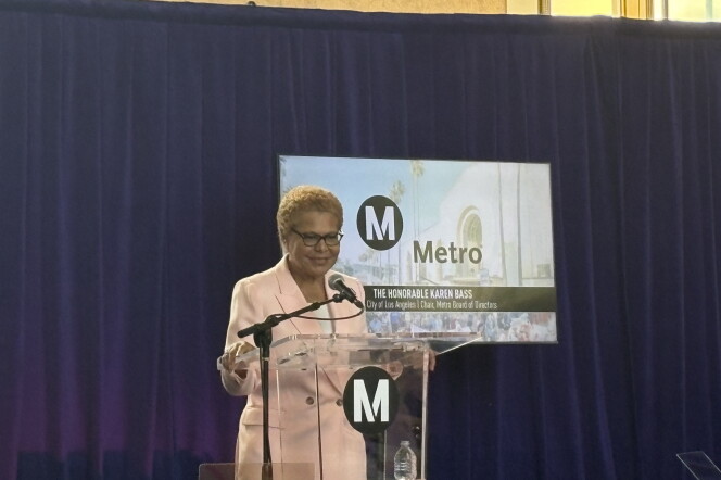 A woman with short brown hair stands in front of a podium with a microphone. The woman is wearing a light pink suit and a white shirt underneath. The podium is emblazoned with the letter "M" in white in a black circle. Behind the woman is a screen that has the same "M" logo and "Metro" written in black. There's a black banner at the bottom of the screen that reads"The Honorable Karen Bass" in all caps.