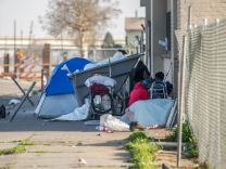 Trash is strewn around an overflowing dumpster on a sidewalk. A blue tent is beside the dumpster.