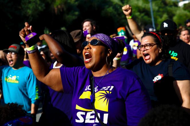 A woman with brown skin, wearing sunglasses, a purple bandanna and a purple t-shirt, shout and raises her fist during a rally. Other protesters are behind her, also with raised fists