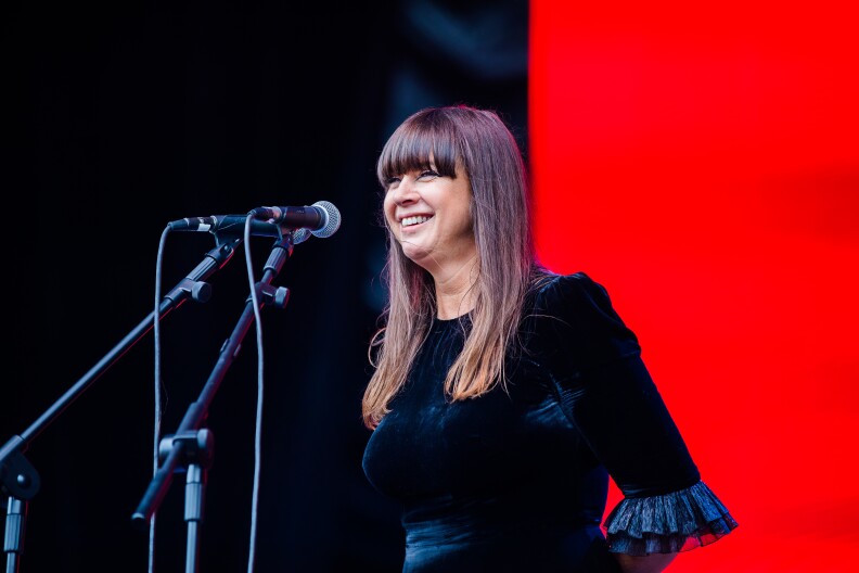 Singer Cat Power stands on stage, smiling, in front of two microphones during the daytime. She's wearing a black velvet dress and has a red background behind her.